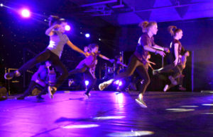 A dance troupe jump in the air on a stage lit by purple lights.