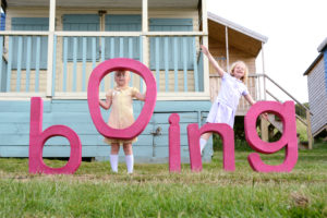 bOing! letters held by 2 little girls in pastel coloured dresses in front of a beach house