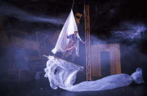 A man is suspended above the stage on a raft with billowing white sheets as sail and sea.