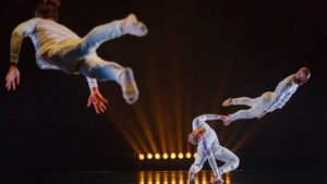 3 dancers in white flyer through the air acrobatically.