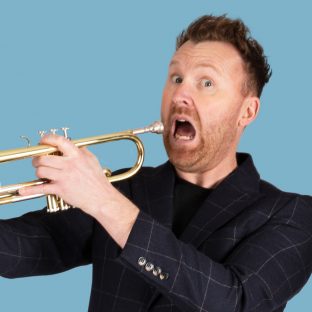 Jason Byrne playing the trumpet while holding a plastic face