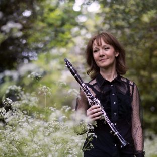 :any holding clarinet, dressed in black blouse and standing in woodland