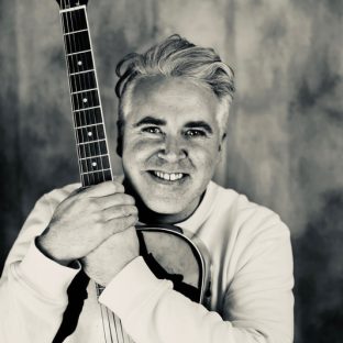 A white middle aged man clutching a guitar and smiling.