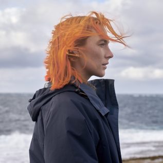 A woman in a blue raincoat with bright orange hair stands in front of the ocean