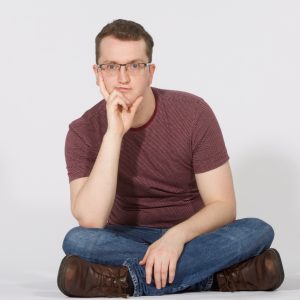 A white man sat crosslegged with his hand on his chin wearing jeans and a burgundy t-shirt